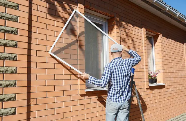 An installer on a ladder fitting a window screen to the side of a brick house